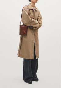 Beżowy oversize trencz z zapinanym przodem, długimi rękawami i bocznym rozcięciem, zestawiony z szerokimi czarnymi spodniami i małą brązową skórzaną torebką.