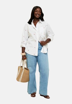 Smiling woman wearing white floral button-up shirt and wide-leg jeans, holding woven tote bag, standing with one hand in pocket and brown sandals.