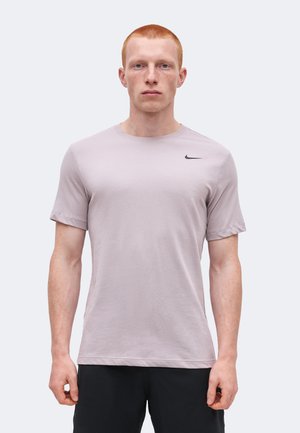 Νέος άνδρας με κοντά κόκκινα μαλλιά, ντυμένος με ανοιχτό μωβ μπλουζάκι Nike και μαύρα σορτς, στέκεται μπροστά σε ένα απλό φωτεινό φόντο.