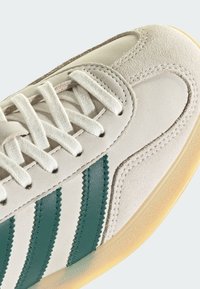 Αθλητικό παπούτσι με λευκό άνω μέρος από σουέτ, τυρκουάζ δερμάτινες ρίγες και υφασμένα διακοσμητικά στοιχεία. Διαθέτει λευκά επίπεδα κορδόνια και ελαφριά ελαστική σόλα.