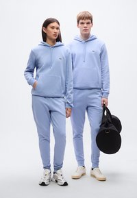 Ανοιχτό γαλάζιο φούτερ με κουκούλα και αντίστοιχο παντελόνι φόρμας από μαλακό ύφασμα, με μπροστινή τσέπη και λογότυπο. Το μοντέλο κρατά μαύρη τσάντα ώμου.