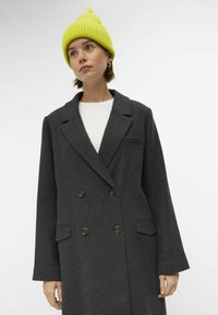 Γκρι διπλής όψης παλτό με δύο μπροστινές τσέπες και μπρούτζινους κουμπιά, φορεμένο πάνω από ένα λευκό τοπ, σε συνδυασμό με ένα φωτεινό κίτρινο πλεκτό σκουφί.