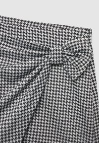 Tkanina w klasyczny wzór houndstooth w kolorach czarnym i białym ma detal w postaci kokardy w talii, o teksturowanym splocie i gładkim wykończeniu.