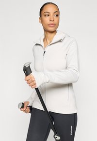 Hellgraue Zip-up-Jacke mit Kapuze aus weichem Stoff; kombiniert mit schwarzen Leggings mit weißem Logo; hält Wanderstöcke.