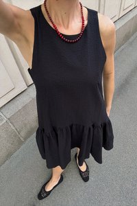 Musta hihaton mekko tekstuurilla ja kerroksellisella röyhelöhelma; yhdistetty mustiin kudottuihin slip-on-kengiin ja punaisiin helmirannekkeisiin.