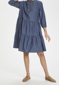 Žena v modrých, dlouhých rukávech a úrovni kolen, šatech s listovým vzorem a knoflíky na hrudi, doplněná hnědými nazouvacími botami.