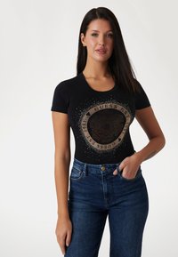 Sort t-shirt med cirkulært metallic grafik og glimmerdetaljer, parret med højtaljede denim jeans. Korte ærmer, rund halsudskæring.