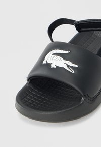 Μαύρο σανδάλι από λείο συνθετικό υλικό, με υφασμένο εσωτερικό πέλμα και λευκό λογότυπο κροκόδειλου σε φαρδιά λουριά. Περιλαμβάνει ρυθμιζόμενο λουρί στο πίσω μέρος.