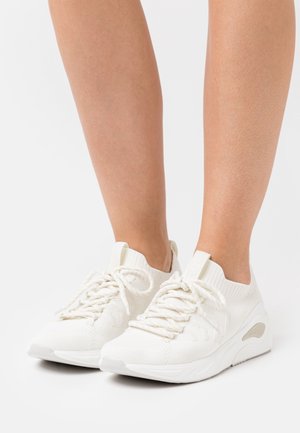 Jambes portant des baskets blanches en tricot avec des lacets et des semelles épaisses, se tenant sur une surface blanche.