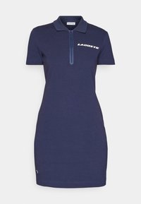 Robe polo manches courtes bleu marine avec fermeture éclair à l’avant et logo Lacoste blanc sur la poitrine.