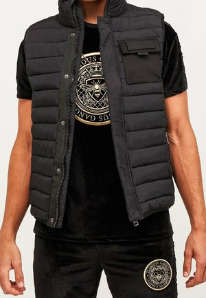Homme portant un gilet matelassé noir sur un t-shirt et un pantalon noirs avec des motifs d'abeille dorée et un emblème circulaire.