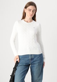 Pull en maille blanche avec des poignets et un ourlet côtelés, arborant un petit logo sur la poitrine. Associé à un pantalon en denim bleu clair.