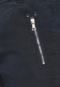 Námořnická modrá tkanina s kapsou na stříbrný zip s hladkým povrchem a kovovými doplňky, umístěná na boku oděvu.