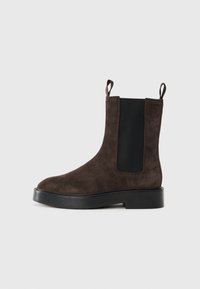 Bottine en suède marron avec des panneaux latéraux élastiques et des languettes de tirage. Présente un bout arrondi et un talon épais noir pour plus de stabilité.