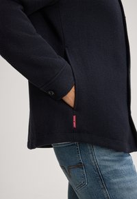 Veste en polaire bleu marine avec poches latérales et poignets à boutons. Équipée d'un label rouge "LOOP JEANS" en bas. Portée avec un jean bleu.