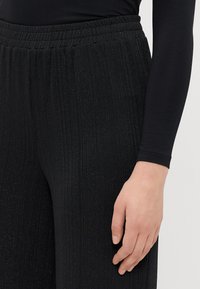 Zwarte, ribbelige broek met een elastische tailleband, met subtiele metallic deeltjes voor extra textuur. Hand rustend op de stof.