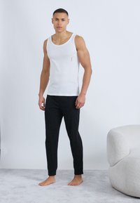 Νεαρός άνδρας όρθιος ξυπόλητος σε χαλί, φοράει λευκό αμάνικο φανελάκι και μαύρο παντελόνι jogger σε έναν μινιμαλιστικό χώρο με λευκούς τοίχους.