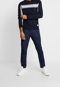 Dunkelblaues Sweatshirt mit weißen Streifen über der Brust, kombiniert mit dunkelblauen gestreiften Hosen. Weiße Sneaker runden das Outfit ab.