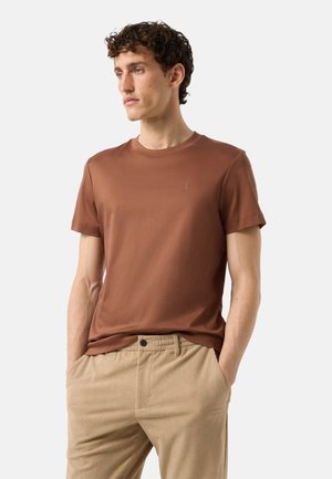 Mand med krøllet hår iført en brun kortærmet t-shirt og beige ribbukser, står med hænderne i lommerne og kigger til venstre.