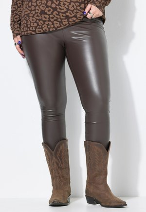 Person trägt enge, dunkelbraune Lederhose, braune bestickte Cowboy-Stiefel und ein langärmeliges Leopardenmuster-Oberteil, mit blauer Nagellackfarbe und Ringen.