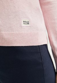 Light pink knit sweater with ribbed hem, featuring a small white tag labeled "POLO CLUB" sewn on the lower left side. Navy pants visible.