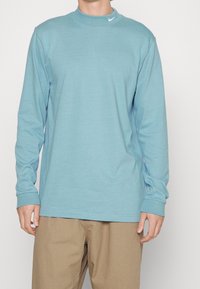 Man i ljusblå långärmad skjorta med liten vit Nike-logga på kragen och beige byxor, stående mot en enhetlig bakgrund.