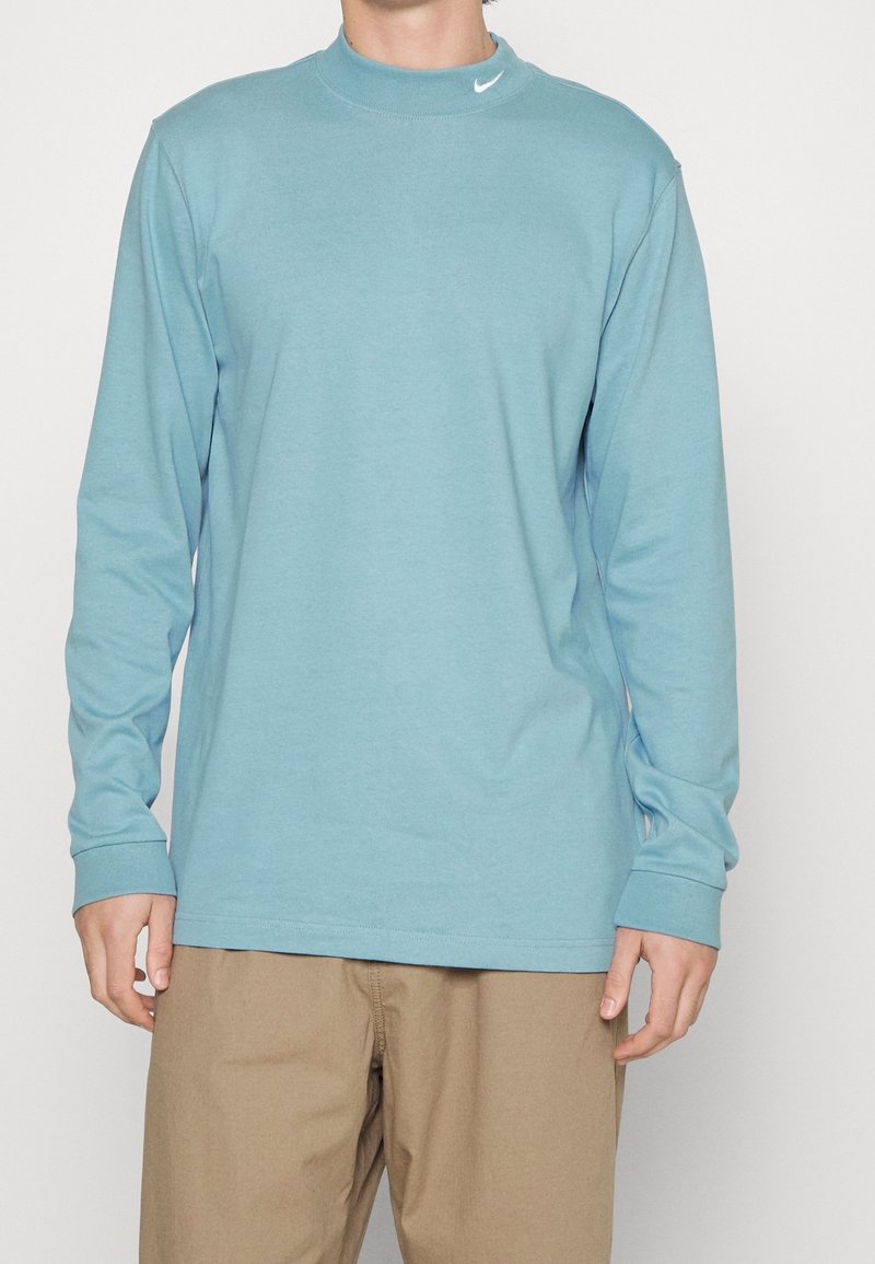 Man i ljusblå långärmad skjorta med liten vit Nike-logga på kragen och beige byxor, stående mot en enhetlig bakgrund.