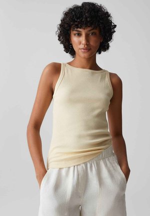 Vrouw met krullend haar draagt een mouwloze beige geribbelde top en lichte broek, staand met handen in de zakken tegen een effen achtergrond.