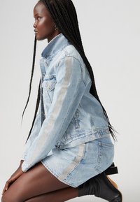 Giacca di jeans azzurro chiaro con strisce verticali argento, dotata di bottoni e tasche, abbinata a una gonna di jeans coordinata e stivaletti neri alla caviglia.