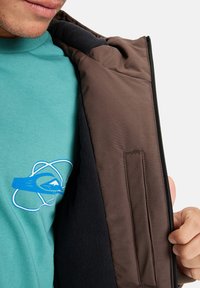 Man som bär en teal-färgad skjorta och som öppnar en brun jacka, vilket visar ett svart fleecefoder och en broderad design på skjortans bröst.