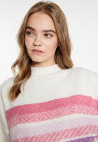 Il maglione presenta una base bianca morbida con strisce orizzontali in rosa, lavanda e viola. Il design include motivi a zigzag variati.