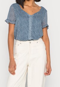 Kortärmad blus i marinblått med vita prickar, v-ringning och mjukt tyg. Matchas med högmidjade byxor i krämfärg.
