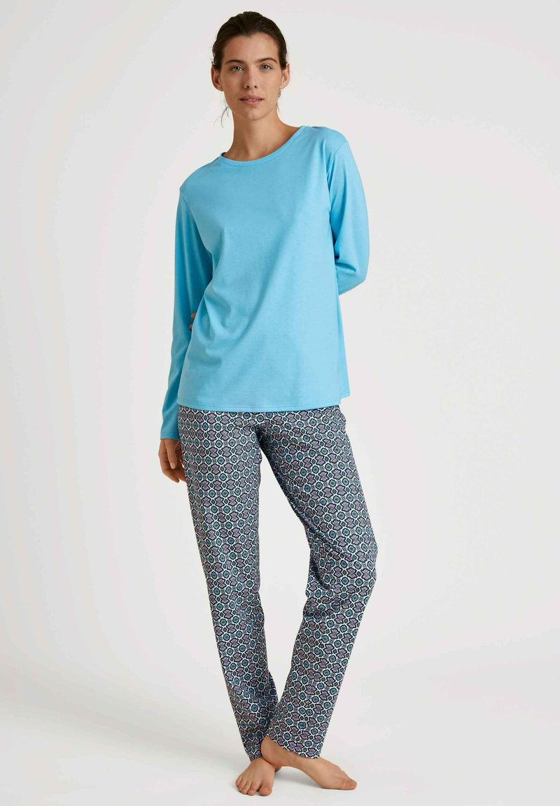 Lichtblauw langemouwtop gecombineerd met een patroonbroek die geometrische ontwerpen in blauw, groen en wit op een donkere achtergrond heeft.