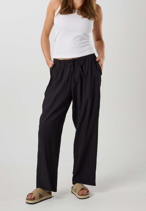 Kobieta w czarnych szerokich spodniach, białym bezrękawnym topie i beżowych sandałach na platformie, stoi z rękami w kieszeniach na jednolitym tle.