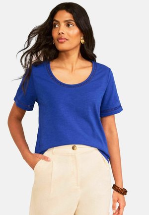 Žena s dlhými tmavými vlasmi, oblečená v modrom tričku s krátkym rukávom a krajkovými detailmi, a krémových nohaviciach s vysokým pásom, pozerajúca sa doľava, s rukou v ruke v náprsnom vrecku.