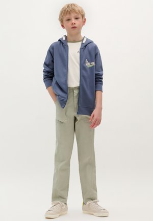 Sudadera azul con cremallera y cordones blancos, texto verde. Camiseta blanca debajo, pantalones verde claro, zapatillas beige, de pie sobre un fondo blanco.