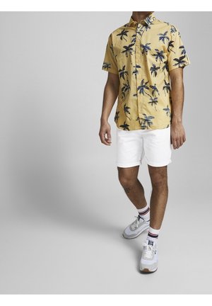 Kurzarm-Bluse in Gelb mit blauem Palmenmuster, kombiniert mit weißen Shorts und Turnschuhen, und gestreiften Socken für zusätzliche Details.