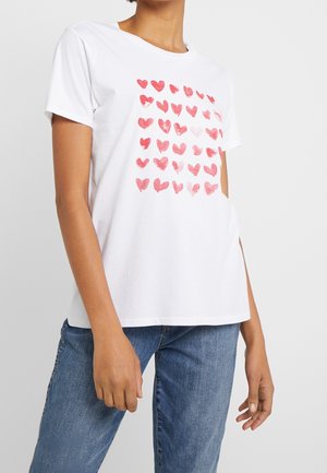 Person iført hvid T-shirt med korte ærmer og et mønster af røde hjerter printet foran, kombineret med blå denimbukser.