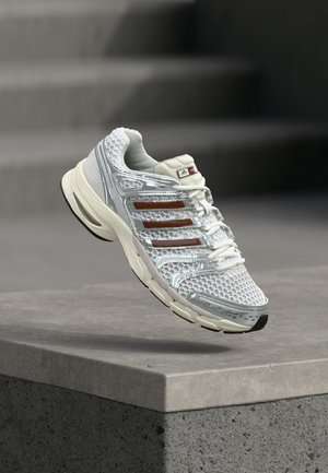 Chaussure de course Adidas blanche et argentée avec des rayures marron flottant au-dessus d'un rebord en béton sur un fond gris flou.