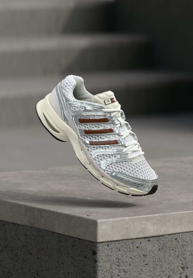 Λευκό και ασημί αθλητικό παπούτσι Adidas με καφέ ρίγες να αιωρείται πάνω από μια τσιμεντένια άκρη με θολό γκρι φόντο.