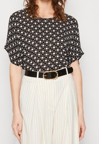 Schwarz-weiß gemusterte lockere Bluse mit kurzen Ärmeln, kombiniert mit weißen Streifenhosen und einem schwarzen Gürtel mit einer goldenen Schnalle.