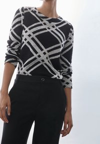 Osoba w czarno-szarym swetrze w kratę i czarnych spodniach na jednolitym jasnym tle.