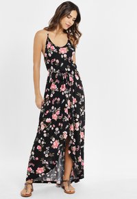Rochie maxi neagră cu flori, cu bretele subțiri, având flori multicolore roz și albe, cu fante laterale și talie ajustabilă printr-un snur. Sandale maro.