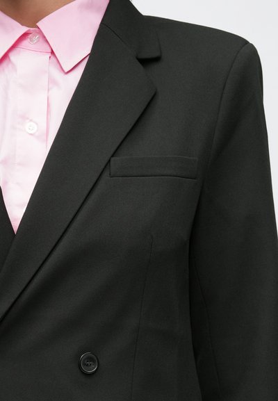 Μαύρο σακάκι με πέτο με εγκοπή, διπλό κούμπωμα με κουμπιά και μία τσέπη στο στήθος, φορεμένο πάνω από ένα ανοιχτό ροζ πουκάμισο με γιακά.