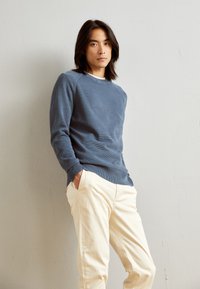 Blau strukturierter Pullover mit gerippten Bündchen, kombiniert mit hellbeigen Hosen. Der Pullover hat einen Rundhalsausschnitt und einen lässigen Schnitt.