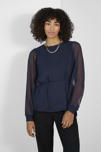 Námořnická modrá blůza s dlouhým rukávem, vyrobená z hladké látky, s nařasenými manžetami a peplum pasem. Průsvitné rukávy dodávají texturu. Kombinováno s černými kalhotami.