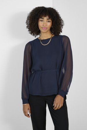 Námořnická modrá blůza s dlouhým rukávem, vyrobená z hladké látky, s nařasenými manžetami a peplum pasem. Průsvitné rukávy dodávají texturu. Kombinováno s černými kalhotami.