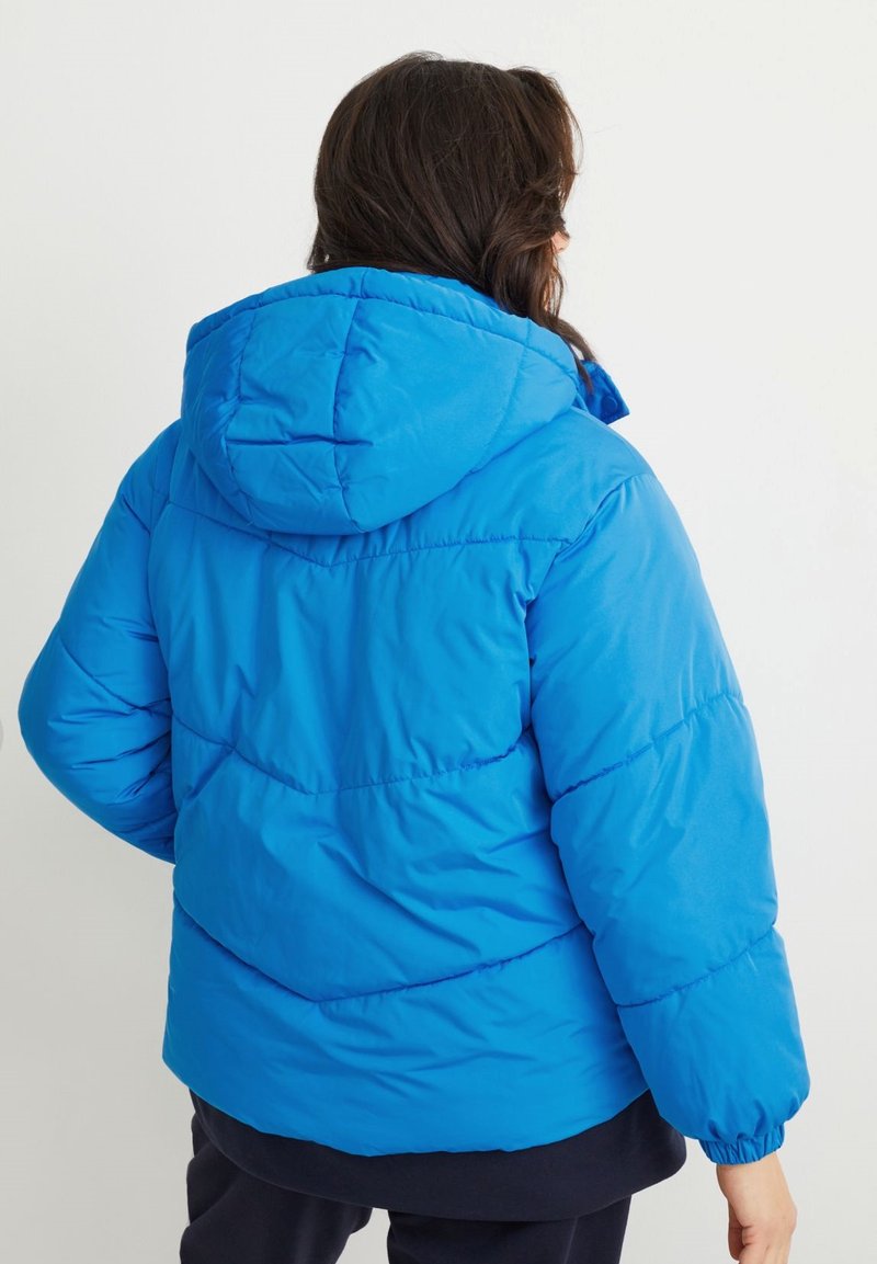 Mujer con el cabello largo y oscuro, wearing una chaqueta acolchada azul brillante con capucha, de espaldas contra un fondo claro y simple.