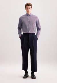 Camicia a righe blu chiaro e bianca; abbinata a pantaloni a pieghe blu scuro. Scarpe eleganti nere, modello in piedi con le mani nelle tasche.