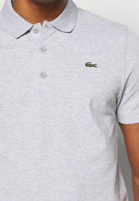 Man i ljusgrå pikétröja med tre knappar och liten grön krokodil-logotyp på vänster bröst.